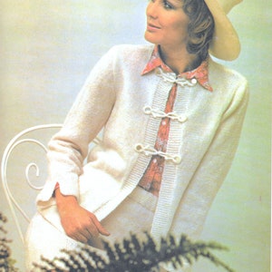 May include: A white knitted cardigan with a white button closure and a white skirt. The cardigan has a textured knit and the skirt has a smooth knit. The model is wearing a pink and white patterned shirt underneath the cardigan. The model is wearing a white hat and is sitting on a white chair.