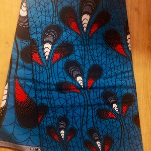 May include: Blue fabric with a repeating pattern of red, white and black stylised peacock feathers. The fabric has the text "BEST QUALITY" and "HOLLAND" printed on the bottom edge.