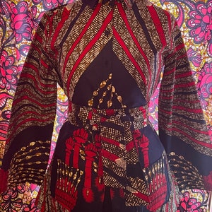 May include: A long-sleeved button-up shirt with a geometric pattern in shades of red, gold, and black. The shirt has a collar and a belt.