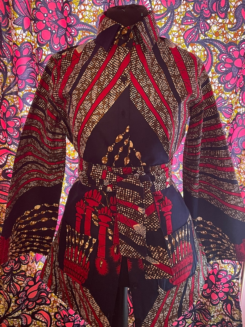 May include: A long-sleeved button-up shirt with a geometric pattern in shades of red, gold, and black. The shirt has a collar and a belt.