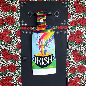 May include: A decorative kitchen towel with the text "Luck of the Irish" and a rainbow design. The towel is hanging on a black, riveted panel. The background features a leopard print with red roses.