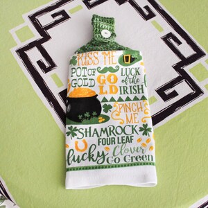 May include: A white kitchen towel with St. Patrick's Day themed text and graphics. The towel features a green crocheted top with a button. Text includes "Kiss Me," "Pot of Gold," and "Shamrock." A black pot of gold is also visible.