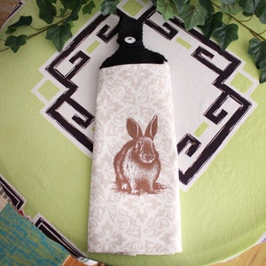 May include: A white kitchen towel with a brown rabbit illustration and a black crocheted hanging loop with a button. The towel has a white and gray floral pattern.