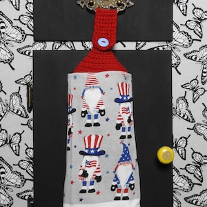 May include: A red crocheted kitchen towel holder with a button closure, holding a gray and white towel. The towel features patriotic gnomes in red, white, and blue. The background is a butterfly pattern.