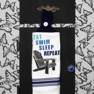 May include: A white kitchen towel with the text "EAT SWIM SLEEP REPEAT" and a graphic of a beach chair. The towel is hanging on a black board with a navy blue crocheted loop and a decorative knob. The background is a gray and white butterfly pattern.