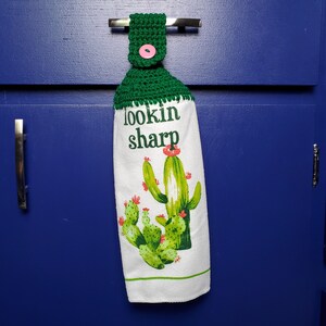May include: A white kitchen towel with a green crocheted hanging loop and a pink button. The towel features a cactus design and the text "lookin sharp".