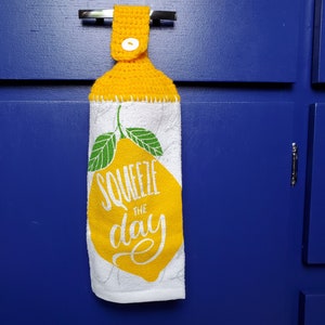 May include: A white kitchen towel with a yellow lemon and the text "Squeeze the day" printed on it. The towel has a yellow crocheted loop at the top for hanging.