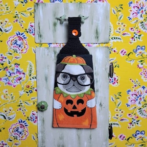 May include: A decorative kitchen towel featuring a pug wearing glasses and a pumpkin costume. The towel hangs from a wooden frame with a green knob and a black hook. The background is a yellow floral pattern.