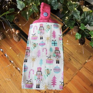 May include: A pink and white kitchen towel with a Christmas nutcracker theme. The towel has a pink crocheted loop with a button and features a repeating pattern of nutcrackers, candy canes, ornaments, and Christmas trees.