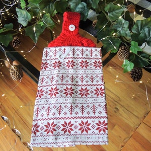 May include: A red and white kitchen towel with a crocheted top. The towel features a repeating pattern of red snowflakes and trees on a white background. A white button secures the crocheted top.