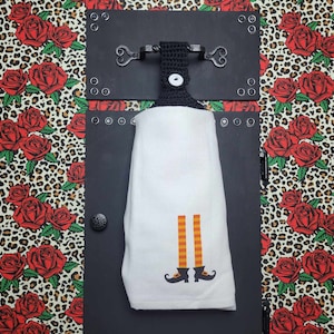 May include: A white kitchen towel with a witch's legs and shoes design. The towel is hanging on a black metal hook with a decorative heart design.