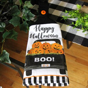 May include: A white kitchen towel with a black and white checkered border. The towel features a black pickup truck filled with orange pumpkins with jack-o-lantern faces. The text "Happy Halloween" and "BOO! 10-31" is printed on the towel.