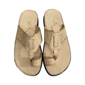 Op de afbeelding: Een paar off-white leren sandalen met ingewikkelde gegraveerde ontwerpen. De sandalen hebben een thong-stijl bandje en een gesloten teen.