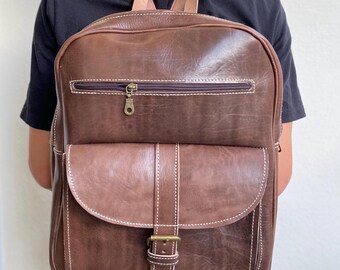 May include: A brown leather backpack with a buckle closure and white stitching. The backpack has a zippered pocket on the front.