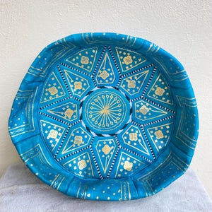 May include: A turquoise blue leather pouf with gold and white geometric patterns. The pouf is round and has a flat top.