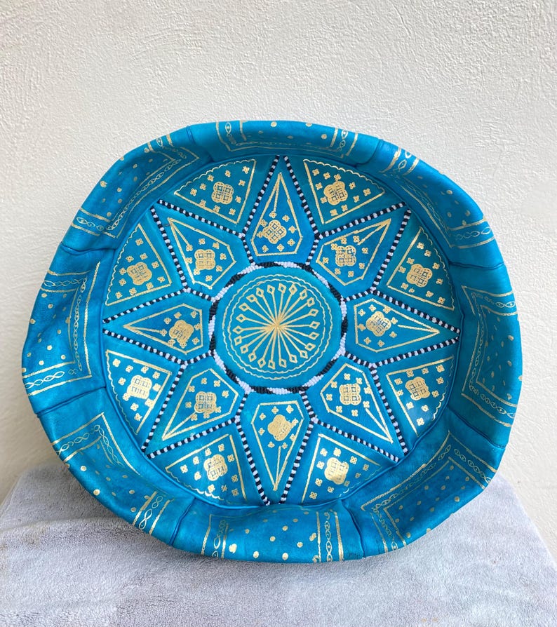 May include: A turquoise blue leather pouf with gold and white geometric patterns. The pouf is round and has a flat top.