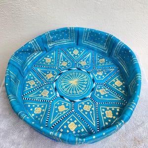 May include: A round, blue leather pouf with gold and white geometric patterns. The pouf is made of leather and has a tufted top.