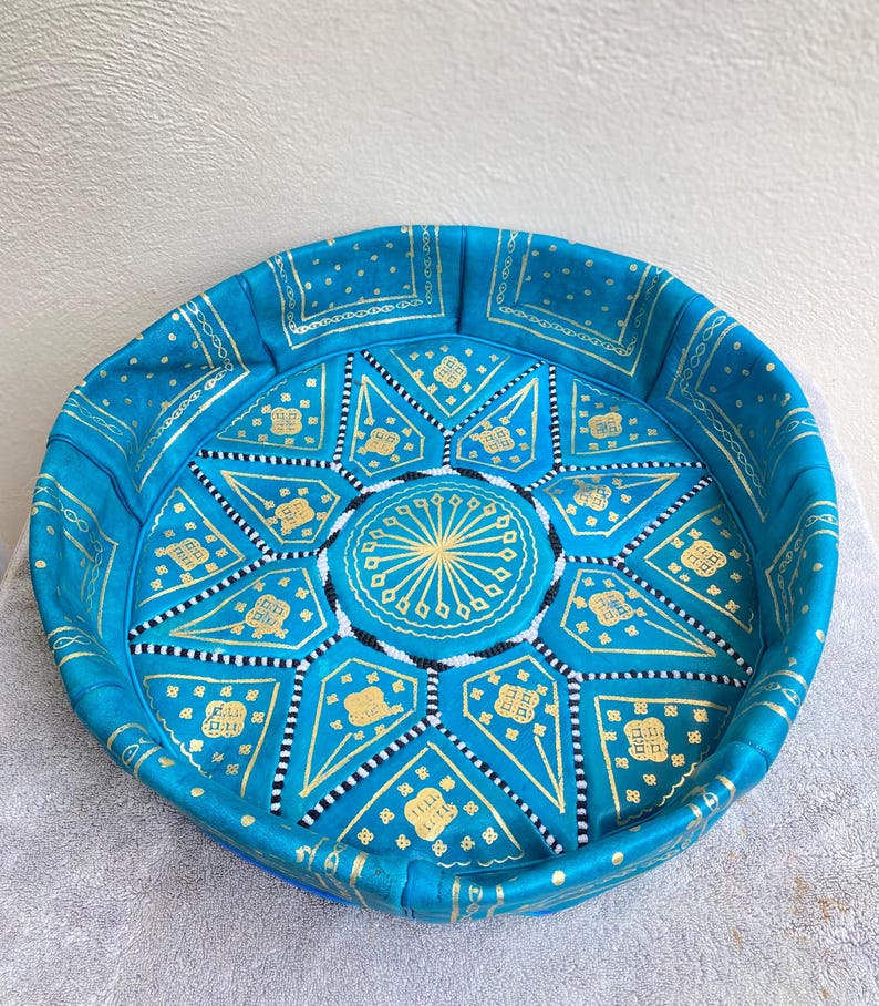 May include: A round, blue leather pouf with gold and white geometric patterns. The pouf is made of leather and has a tufted top.