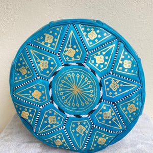 May include: A round turquoise leather pouf with a gold and white geometric pattern. The pouf is decorated with small white beads.