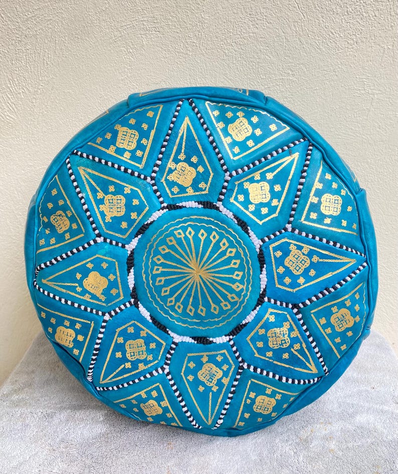 May include: A round turquoise leather pouf with a gold and white geometric pattern. The pouf is decorated with small white beads.