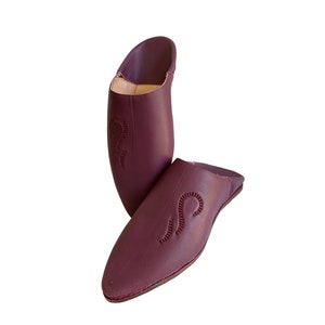 May include: A pair of burgundy leather slippers with intricate embroidered designs. The slippers have a pointed toe and a flat sole.