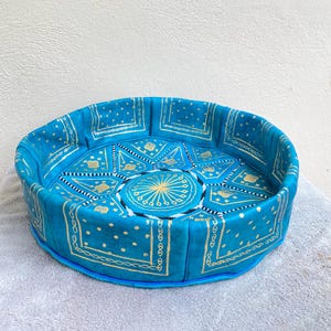 May include: A round, blue leather pouf with a gold and white geometric pattern. The pouf is made of leather and has a fringed edge.