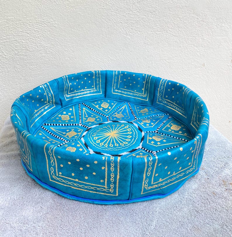 May include: A round, blue leather pouf with a gold and white geometric pattern. The pouf is made of leather and has a fringed edge.
