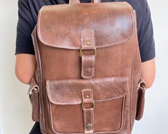 May include: A brown leather backpack with two front pockets and a buckle closure. The backpack has white stitching and gold-toned hardware.