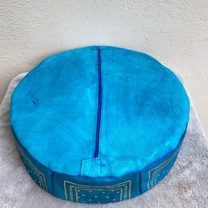 May include: A round blue leather pouf with a zipper closure and a gold bandana pattern around the bottom.