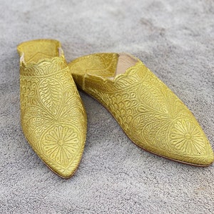 Puede incluir: Un par de zapatillas bordadas doradas con punta puntiaguda. Las zapatillas tienen un patrón floral y están hechas de una tela suave y lujosa.