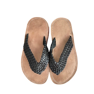 Könnte beinhalten: Ein Paar braune Ledersandalen mit schwarzen geflochtenen Riemen. Die Sandalen haben ein schlichtes Design und sind perfekt für warmes Wetter.