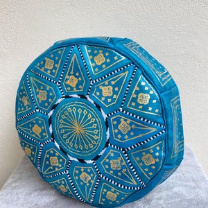May include: A turquoise blue leather pouf with a gold and white geometric pattern. The pouf is round and has a flat top.