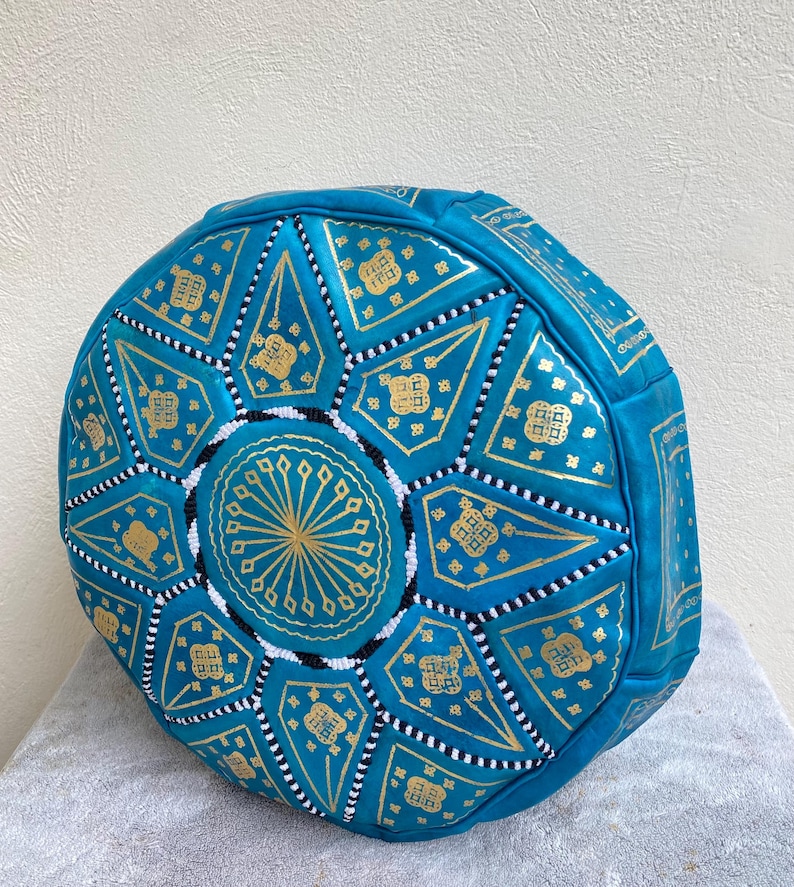 May include: A turquoise blue leather pouf with a gold and white geometric pattern. The pouf is round and has a flat top.