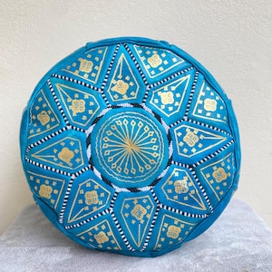 May include: A turquoise blue leather pouf with a gold and white geometric pattern. The pouf is round and has a flat top.