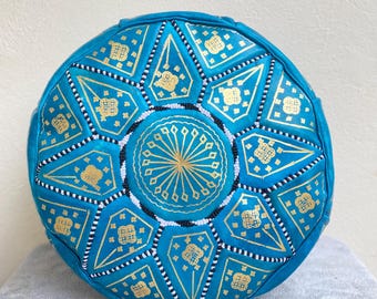 May include: A turquoise blue leather pouf with a gold and white geometric pattern. The pouf is round and has a flat top.