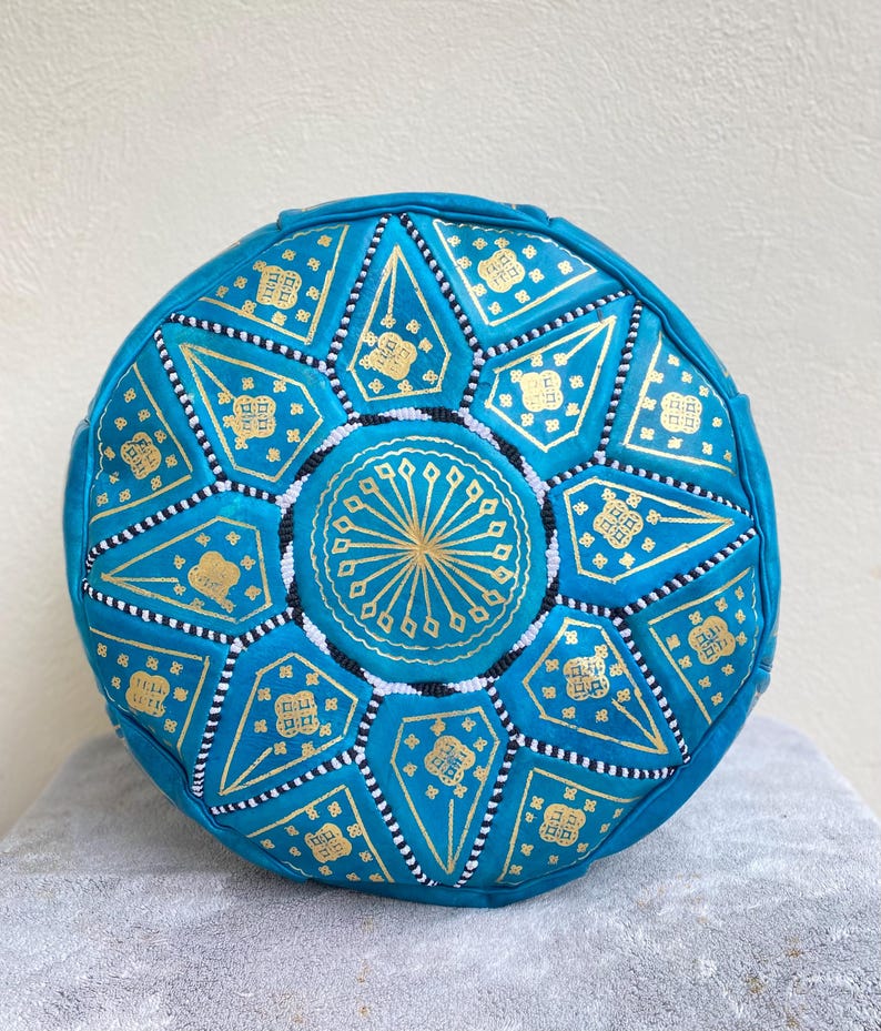 May include: A turquoise blue leather pouf with a gold and white geometric pattern. The pouf is round and has a flat top.