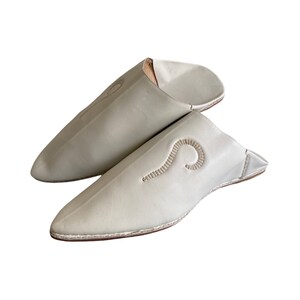 May include: A pair of white leather slippers with a pointed toe and a simple embroidered design. The slippers are made of soft, supple leather and have a comfortable fit.