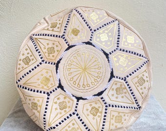 May include: A cream-colored leather pouf with a geometric pattern in gold and black. The pouf is round and has a tufted top.