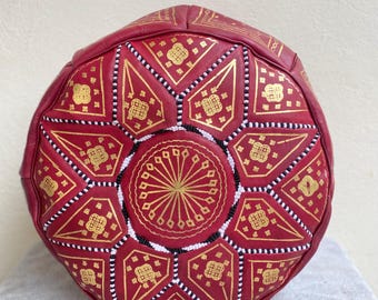 May include: A round, red leather pouf with gold geometric patterns. The pouf features a central sunburst design and is adorned with gold accents and white and black beaded trim. This decorative piece adds a touch of bohemian flair to any living space.