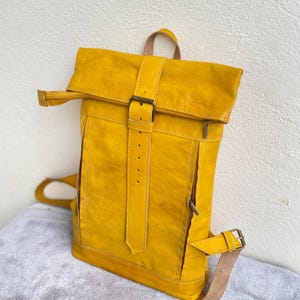 Könnte beinhalten: Ein gelber Lederrucksack mit einer umklappbaren Klappe und einem Schnallenverschluss. Der Rucksack hat eine Reißverschlusstasche vorne und verstellbare Träger.