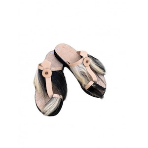 Könnte beinhalten: Ein Paar handgefertigte Sandalen mit einzigartigem Design. Die Sandalen haben eine beige Lederbasis, schwarze Sohlen und dekorative Fellakzente in Schwarz, Weiß und Hellbraun. Eine kleine, runde, hellbraune Lederblume verziert die Riemen.
