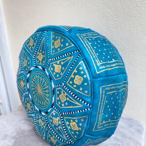 May include: A turquoise leather pouf with gold geometric patterns. The pouf is round and has a tufted design.