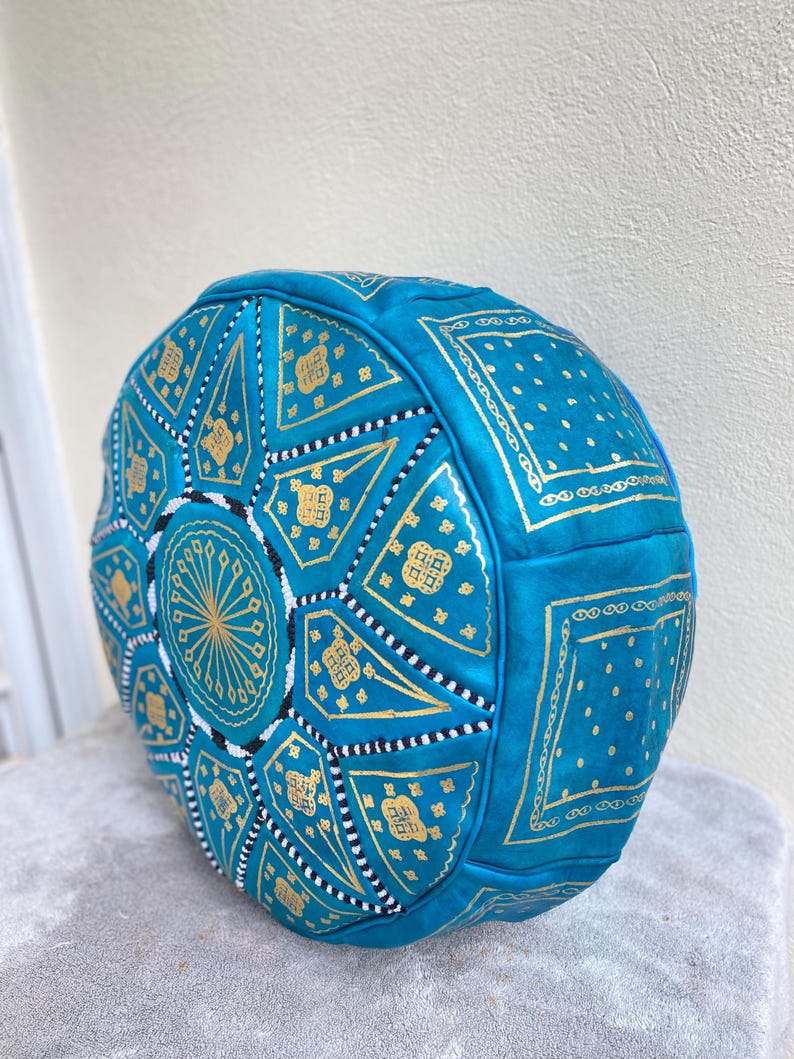 May include: A turquoise leather pouf with gold geometric patterns. The pouf is round and has a tufted design.