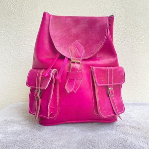 May include: A bright pink leather backpack with two front pockets and a buckle closure. The backpack has a drawstring closure at the top.