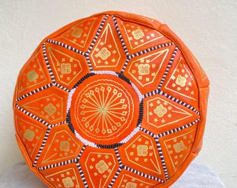 May include: Orange leather pouf with a geometric pattern in gold and white. The pouf is round and has a tufted top.