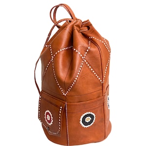 May include: A brown leather drawstring backpack with white stitching and decorative embroidery. The backpack has a large main compartment and a smaller front pocket.
