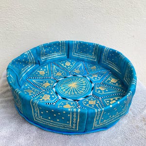 May include: A round, blue leather pouf with a gold and white geometric pattern. The pouf is made of leather and has a fringed edge.