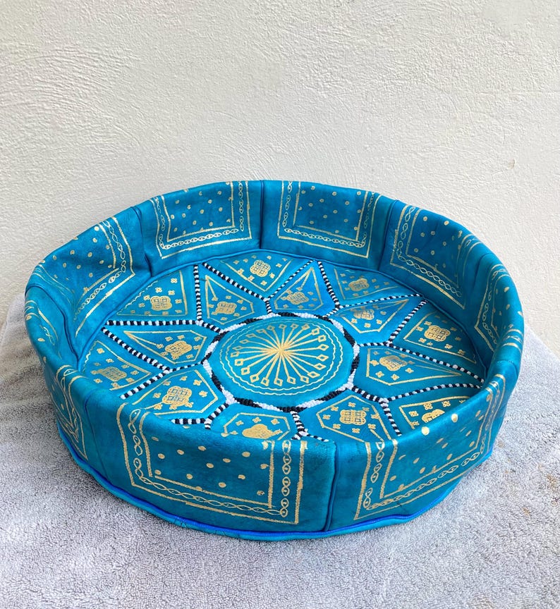 May include: A round, blue leather pouf with a gold and white geometric pattern. The pouf is made of leather and has a fringed edge.