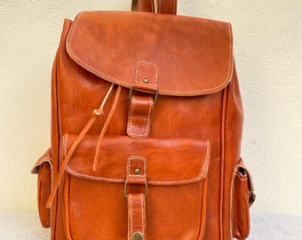 May include: A brown leather backpack with a flap closure and multiple pockets. The backpack has a buckle and a strap with a tassel.