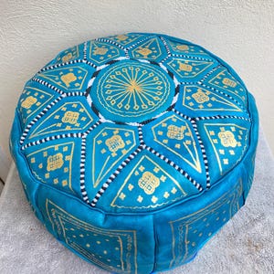 May include: A turquoise blue leather pouf with gold and white geometric patterns. The pouf is round and has a tufted top.